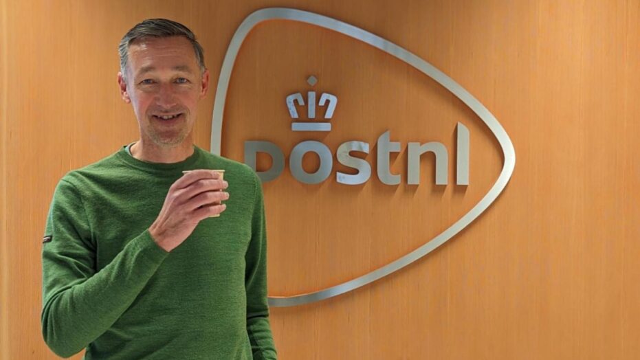 Marcel van Rooijen staat voor het logo van PostNL met een kopje koffie in zijn hand